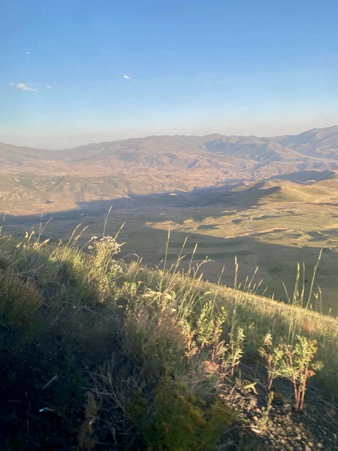 View from Vayots Sar across sunlit and shadowed ridges of Vayots Dzor and the Arpa Valley, with grasses on the summit slope in the foreground.