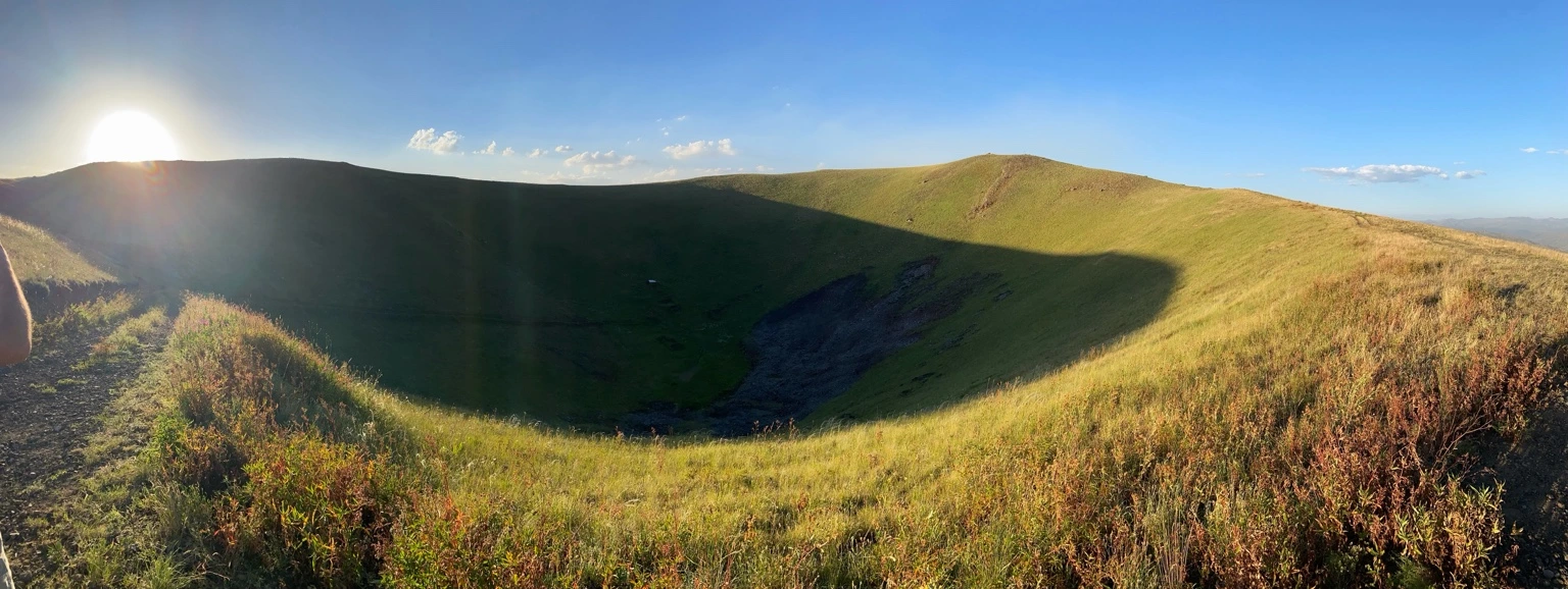 Wide view from the rim into the grassy bowl of Vayots Sar, with low sunlight crossing the crater and a shadow over its floor.