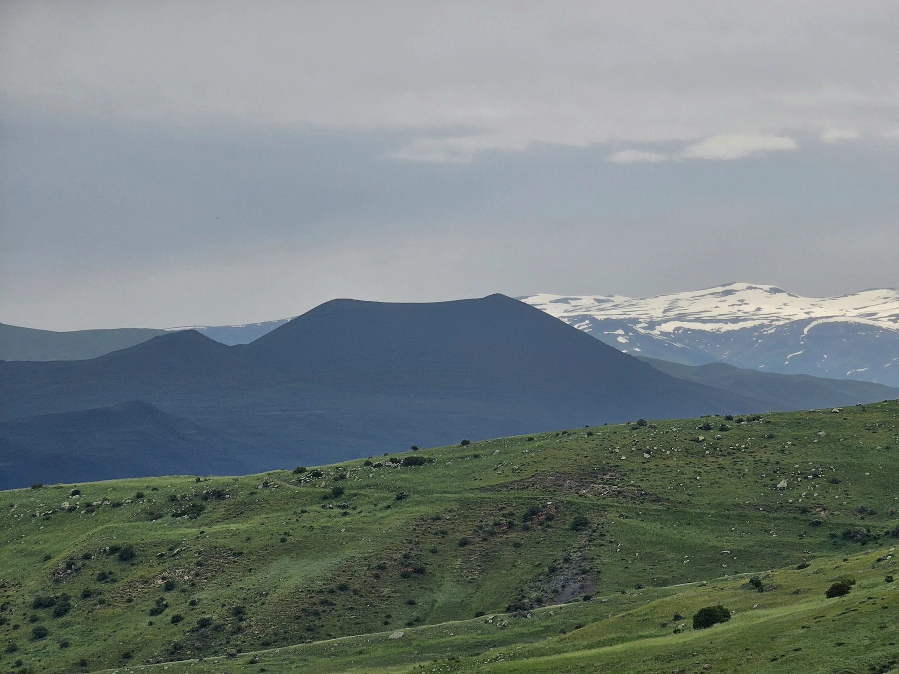 The flat-topped silhouette of Vayots Sar beyond green hills, with snowy mountains in the distance.
